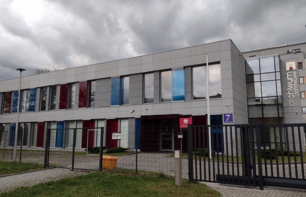 Budynek gorzowskiego Archiwum. Na pierwszym planie zamknięta brama. Nad archiwum ciemne, burzowe chmury. 