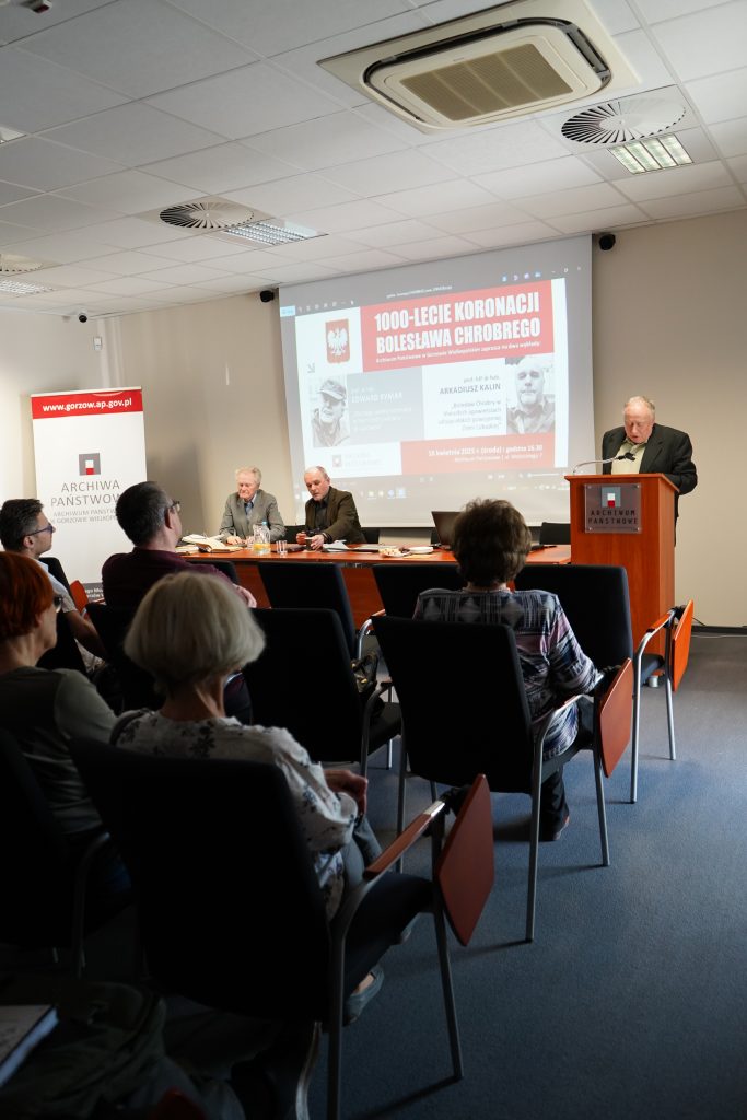 W sali konferencyjnej za stołem prezydialnym siedzą dwaj mężczyźni. Trzeci przemawia za pulpitem. Za nimi wyświetlony jest slajd. Na pierwszym planie kilka osób siedzących na krzesłach i zwróconych w stronę przemawiających. 