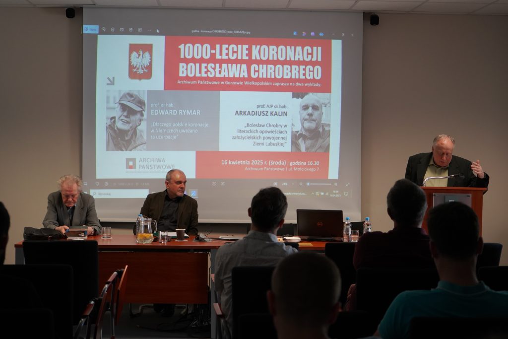 W sali konferencyjnej za stołem prezydialnym siedzą dwaj mężczyźni. Trzeci przemawia za pulpitem. Za nimi wyświetlony jest slajd. Na pierwszym planie kilka osób siedzących na krzesłach i zwróconych w stronę przemawiających. 