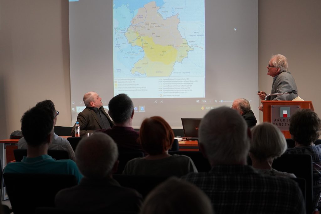 W sali konferencyjnej za stołem prezydialnym siedzi mężczyzna. Drugi mężczyzna przemawia za pulpitem i pokazuje na mapę wyświetloną na slajdzie. Na pierwszym planie kilkanaście osób siedzących na krzesłach i zwróconych w stronę przemawiających. 