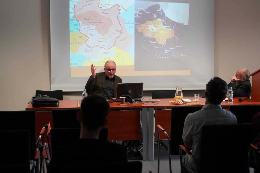 W sali konferencyjnej za stołem prezydialnym siedzą dwaj mężczyźni. Jeden z  nich przemawia Za nimi wyświetlony jest slajd. Na pierwszym planie kilka osób siedzących na krzesłach i zwróconych w stronę przemawiających. 