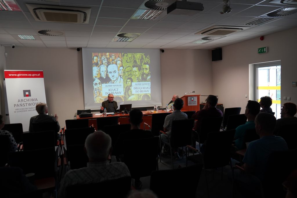 W sali konferencyjnej za stołem prezydialnym siedzą dwaj mężczyźni. Jeden z nich przemawia Za nimi wyświetlony jest slajd. Na pierwszym planie kilkanaście osób siedzących na krzesłach i zwróconych w stronę przemawiających. 
