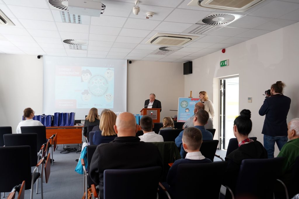 W sali konferencyjnej kilkanaście osób siedzi na krzesłach. Zwróceni są w stronę przemawiającego mężczyzny. Obok niego stoi kobieta. Przy ścianie stoi mężczyzna i fotografuje przemawiającego.