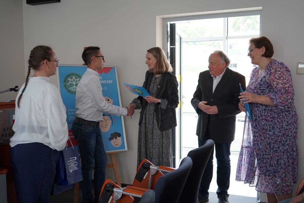 Kobieta wręcza dyplom finaliście konkursu. Obok niej stoi mężczyzna i dwie kobiety. 