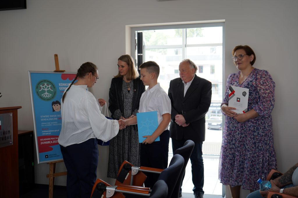 Kobieta wręcza dyplom finaliście konkursu. Obok niej stoi mężczyzna i dwie kobiety. 