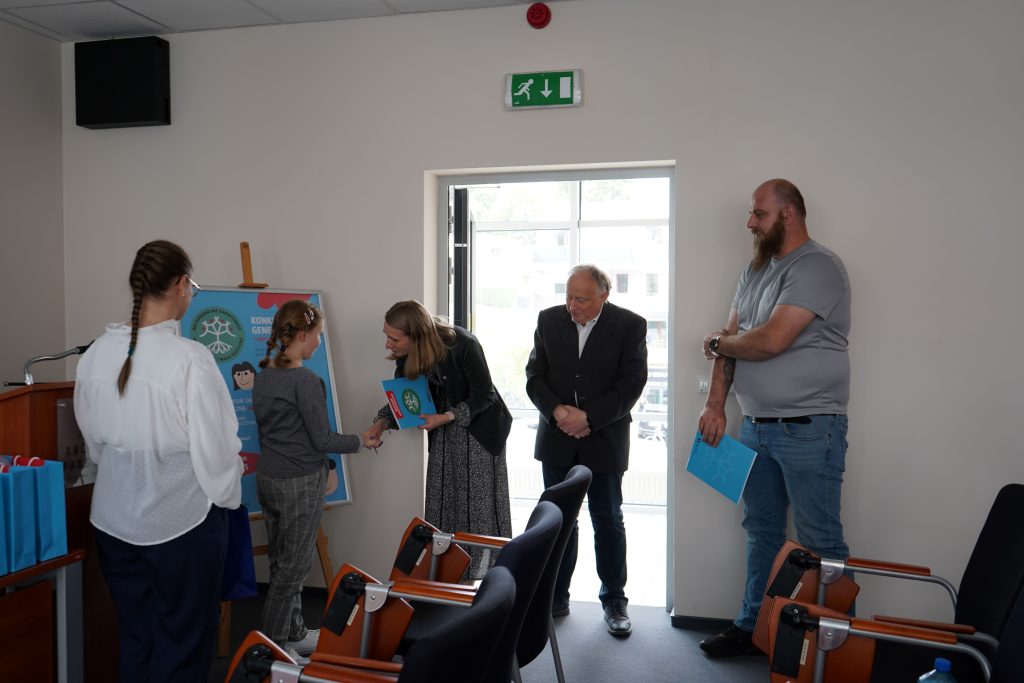 Kobieta wręcza dyplom finalistce konkursu. Obok niej stoi dwóch mężczyzn.