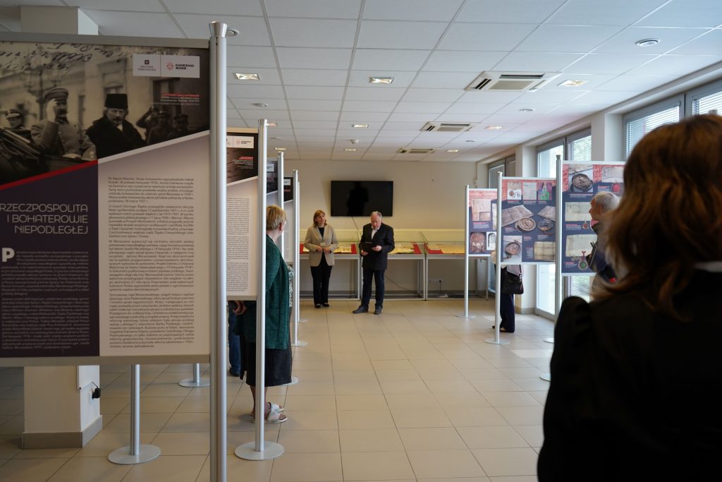 Dwie osoby przemawiają podczas otwarcia wystawy. Za nimi widać gabloty z aktami. Po obu stronach stoją stojaki z planszami na stojakach.