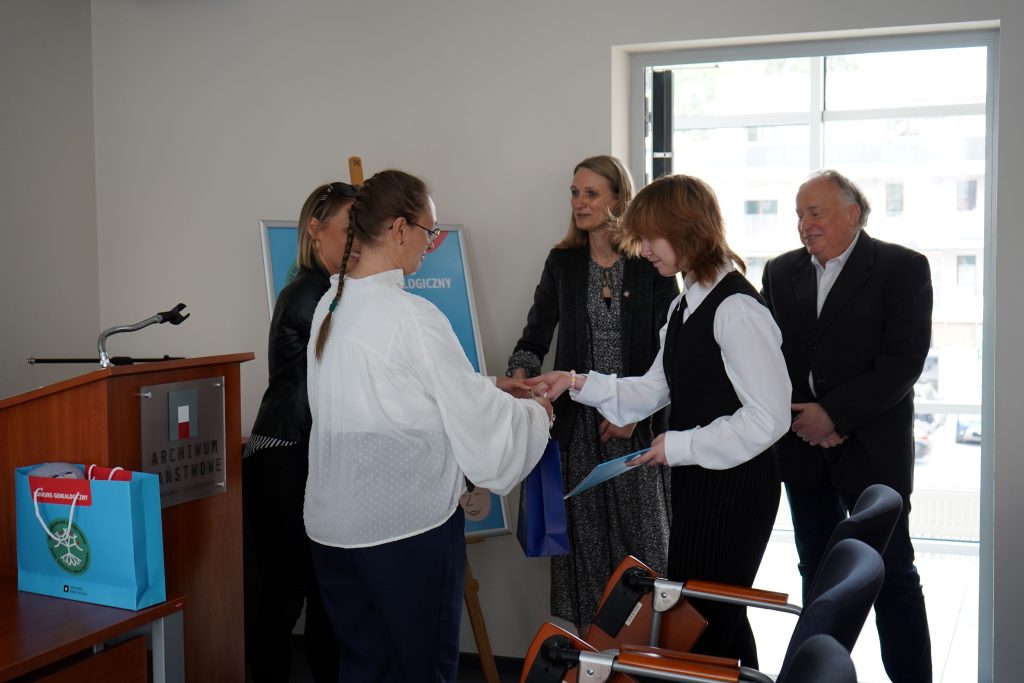 Kobieta wręcza dyplom finalistce konkursu. Obok niej stoi mężczyzna i dwie kobiety. 