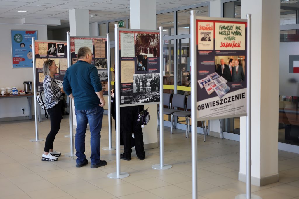 W sali wystawowej kilka osób ogląd a wystawę na stojakach.