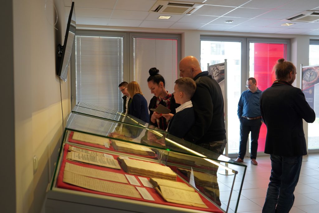 Kilka osób ogląda księgi zamieszczone w podświetlanych gablotach. Kilka osób ogląda wystawę na planszach. 