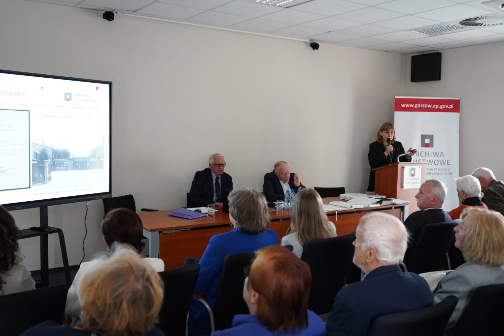 Spotkanie w sali konferencyjnej. Kobieta przemawia do zgromadzonych gości.