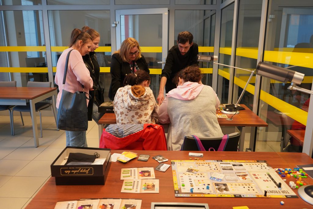 W czytelni prezentacja gry planszowej. Kilka kobiet i mężczyzna prowadzą rozgrywaki.