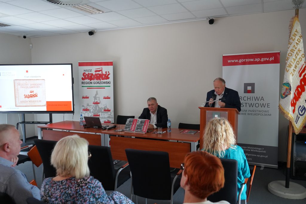 Dyrektor Archiwum przemawia do gości. Obok siedzi drugi mężczyzna. Po lewej stronie widać ekran z wyświetlonym sztandarem. Po prawej stronie stoi sztandar. 