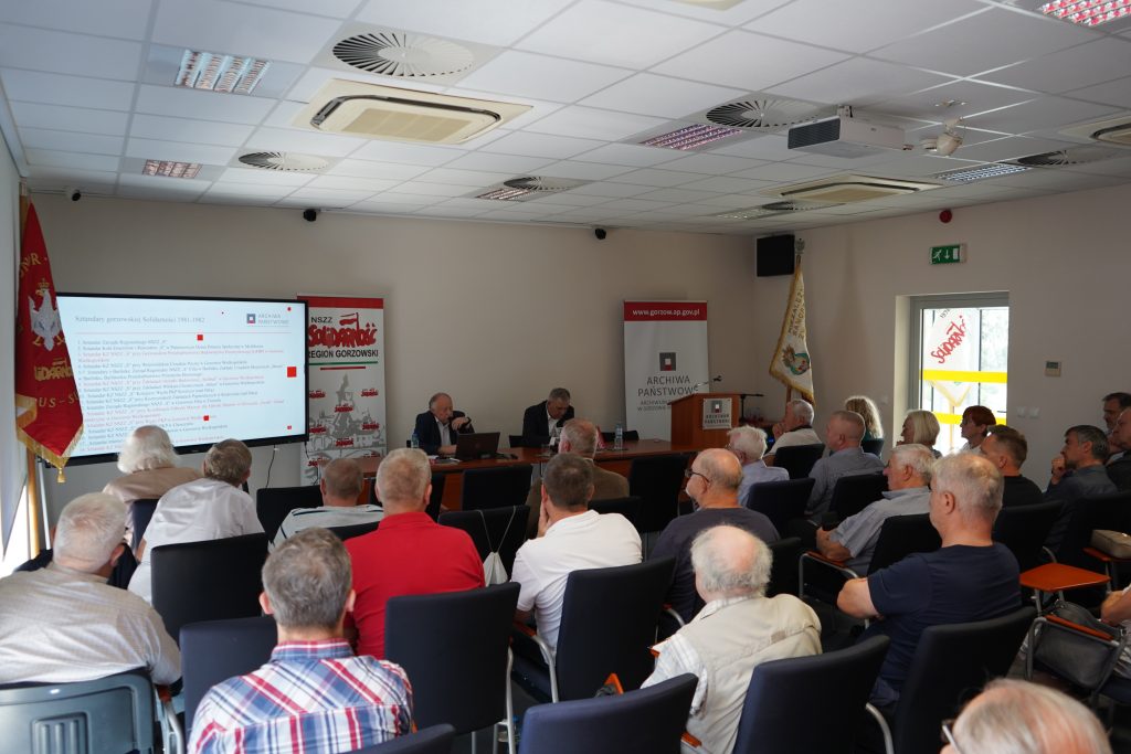 Widok na salę konferencyjną, w której odbywa się promocja książki. Z tyłu widać przemawiającego dyrektora archiwum