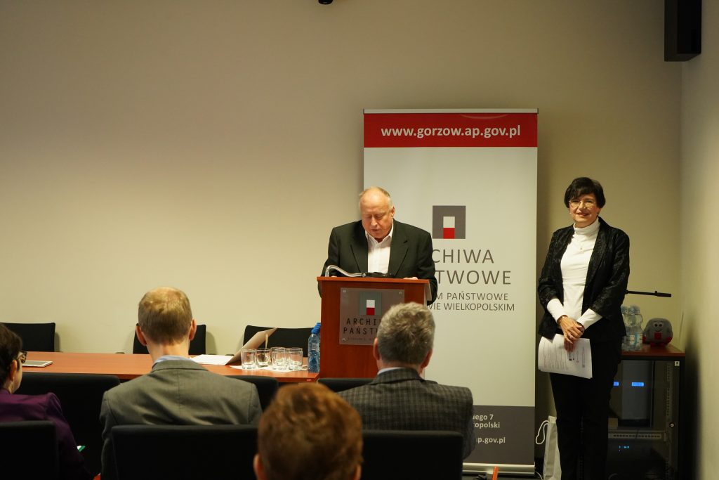 Sala konferencyjna. Mężczyzna przemawia za pulpitem, obok stoi kobita.