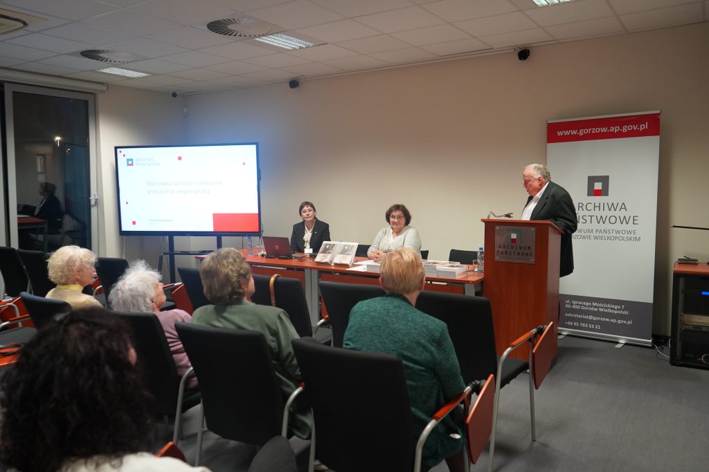 Promocja książki w sali konferencyjnej. Za stołem siedzą dwie kobiety. Mężczyzna przemawia przy pulpicie. 